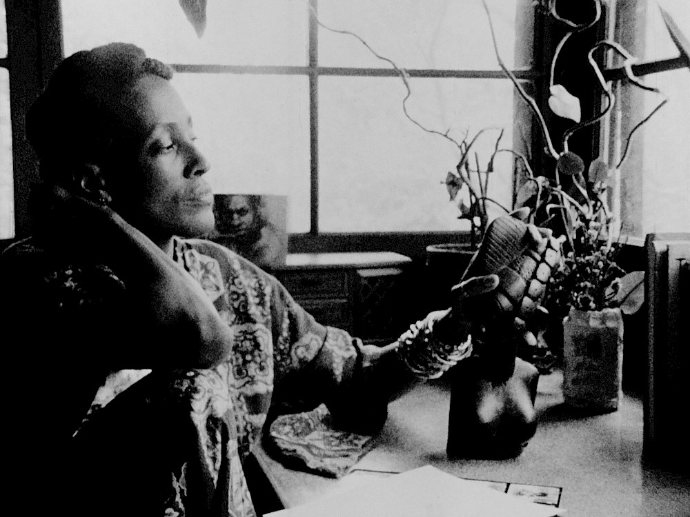 A black woman is sat down at a domestic desk by a window, with one hand resting behind her head and her other hand caressing a small sculpture of a female bust.