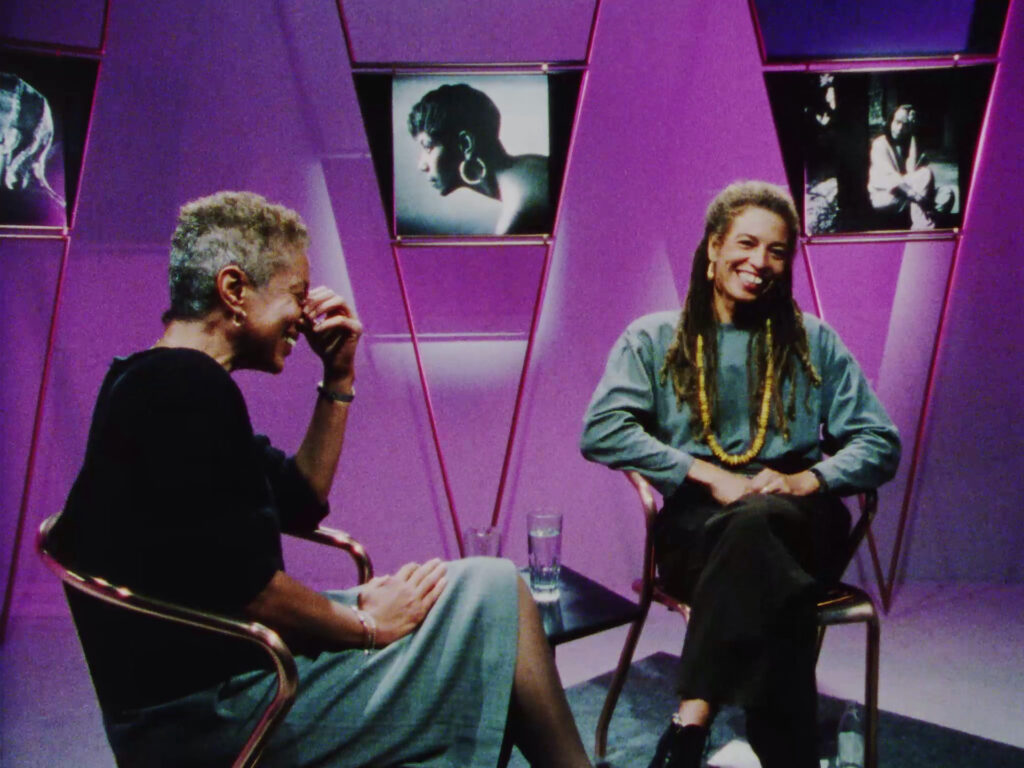 Two Black women, June Jordan and Angela Davis are sitting closely together in a studio installation, with purple walls, and black and white photographs on long triangular stands. Angela Davis is smiling and looking at the camera, while June Jordan is laughing and smiling with her eyes closed and one hand up to her face.